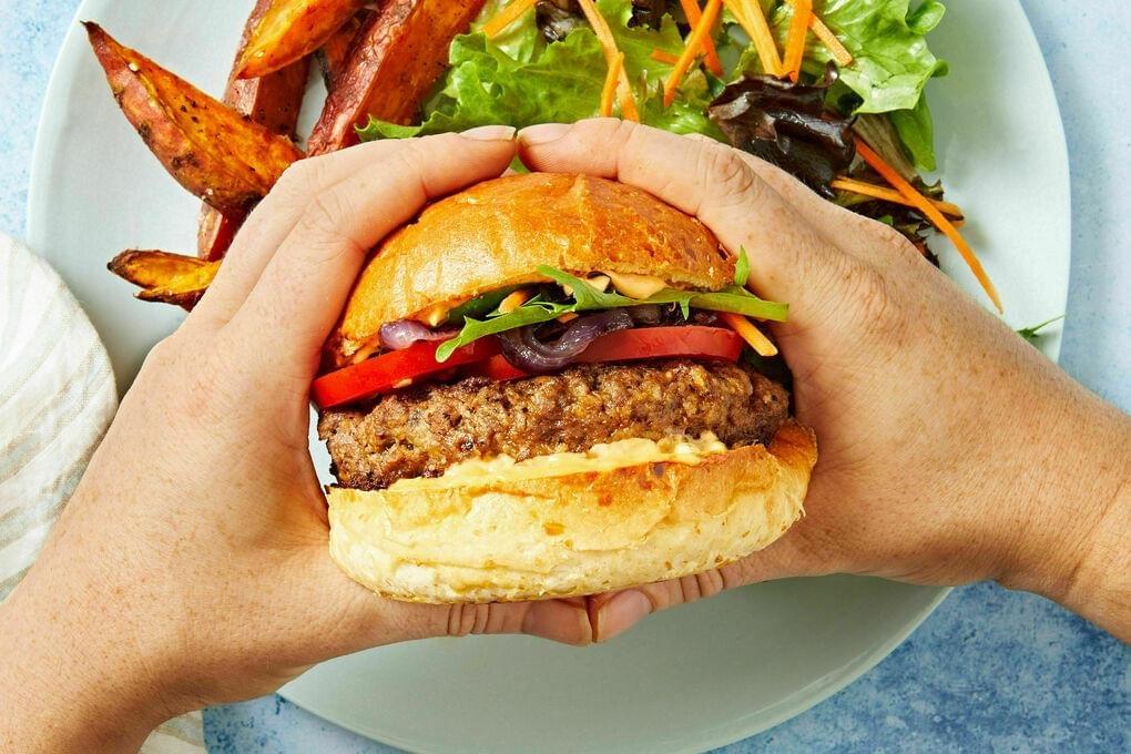 A person holds a thick, toasted burger layered with tomato, red onion, and fresh greens between their hands. The meal is served on a white plate alongside roasted sweet potato wedges and a crisp side salad.