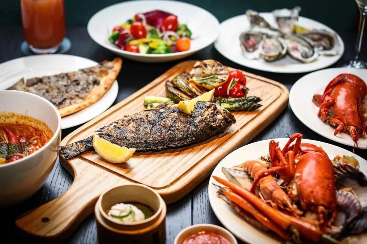 A wooden cutting board with a whole grilled fish, lemon slices, and roasted vegetables sits at the center of a dark table filled with various dishes. Surrounding the central board are plates featuring fresh oysters, cooked lobsters, flatbread, a vibrant garden salad, and bowls of dipping sauces.