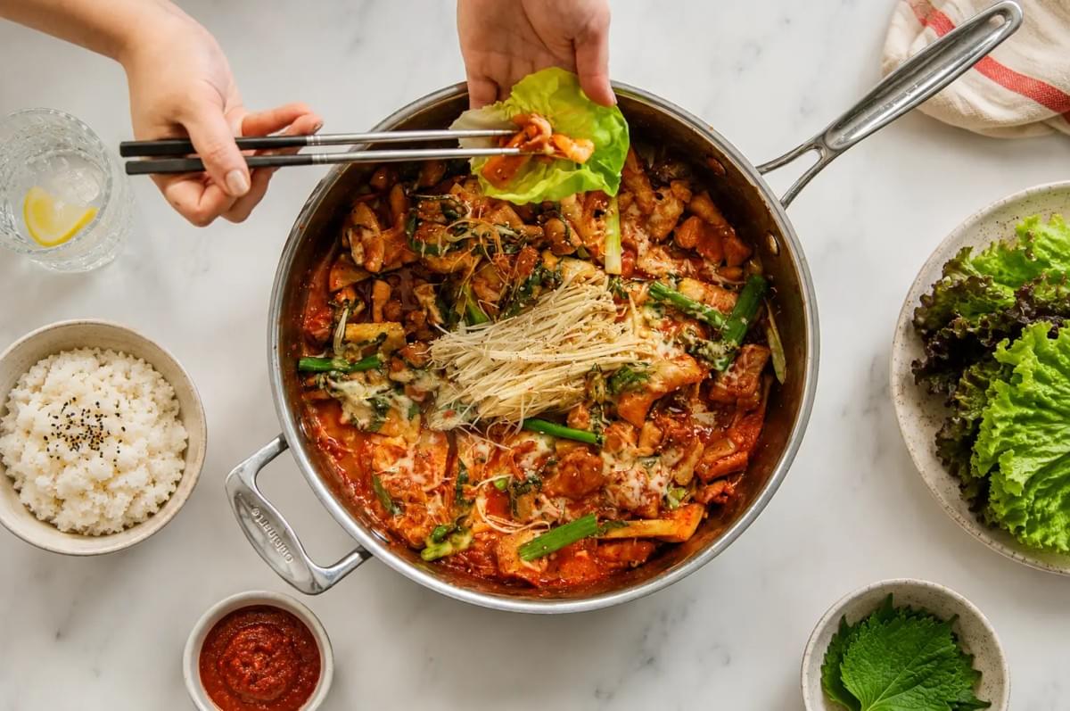 An overhead view of a large stainless steel pan filled with cheesy dak-galbi and enoki mushrooms, as hands use chopsticks to assemble a lettuce wrap, surrounded by bowls of rice and side dishes.