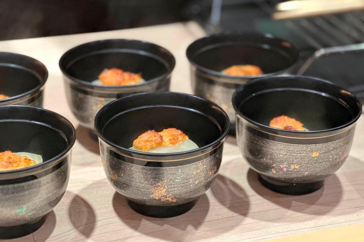 A close-up shot of six elegant black lacquer bowls with gold and iridescent flecked detailing, arranged on a light wood surface. Inside each bowl is a single, refined dish: a pale, translucent slice of vegetable (possibly simmered daikon) topped with a dollop of toasted or seared orange garnish, likely sea urchin (uni) or a savory miso paste. The lighting is soft, highlighting the texture of the food.