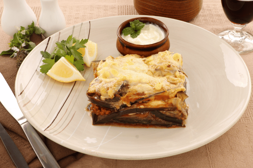 A golden slice of Greek moussaka layered with tender eggplant, spiced ground meat, and creamy béchamel, served with lemon wedges, parsley, and mint-topped yogurt sauce on a white plate—evoking warmth, richness, and Mediterranean comfort.