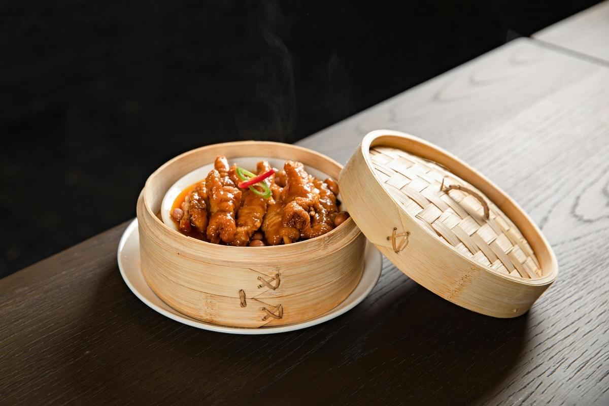 A steaming dish of braised dim sum chicken claws is served in a small, round plate inside an open bamboo steamer basket on a dark wooden table. The woven bamboo lid rests angled against the side of the basket, revealing the glossy, sauce-coated dish garnished with a slice of chili.