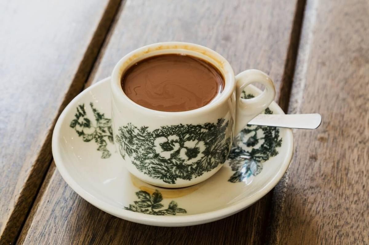 A rich, brown cup of coffee in a small, white ceramic cup with floral green patterns, sits on a matching saucer with a spoon, atop a wooden table.