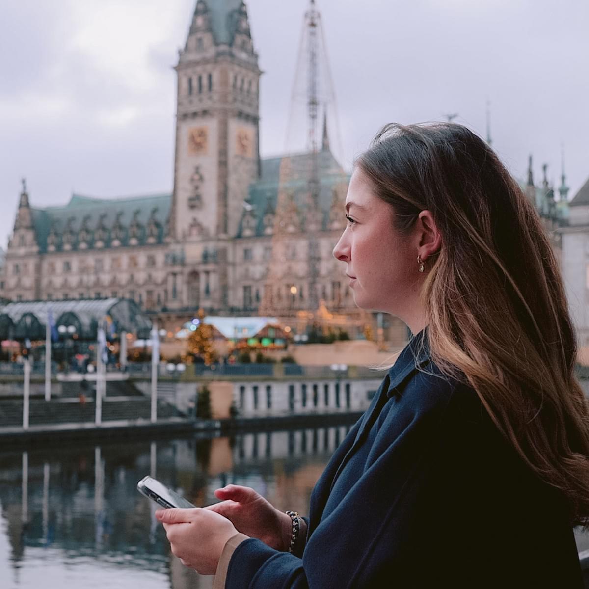 Eine junge Frau mit Handy in der Hand guckt in die Ferne, im Hintergrund das Hamburger Rathaus