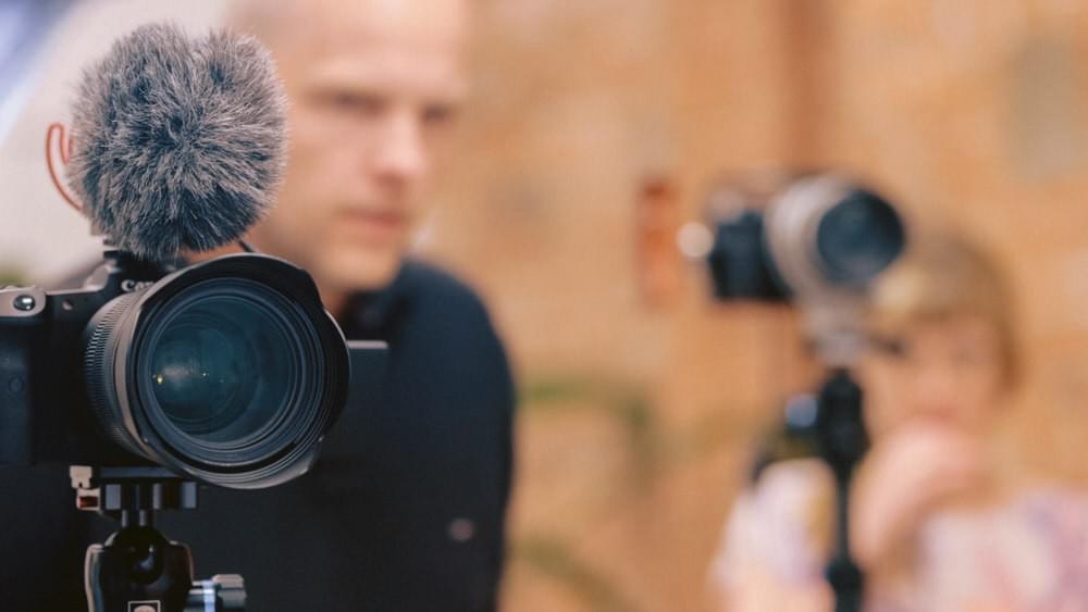 Bild von einer Produktion. Eine Kamera scharf im Vordergrund, zwei Personen und eine Person unscharf im Hintergrund.