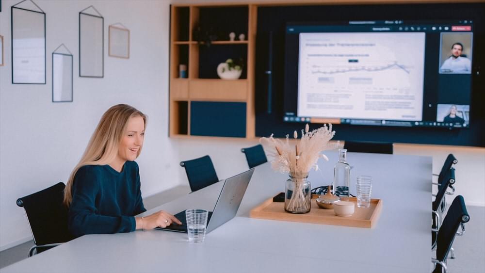 Frau in einem Meetingraum spricht im Online-Call am Laptop mit einem Kollegen