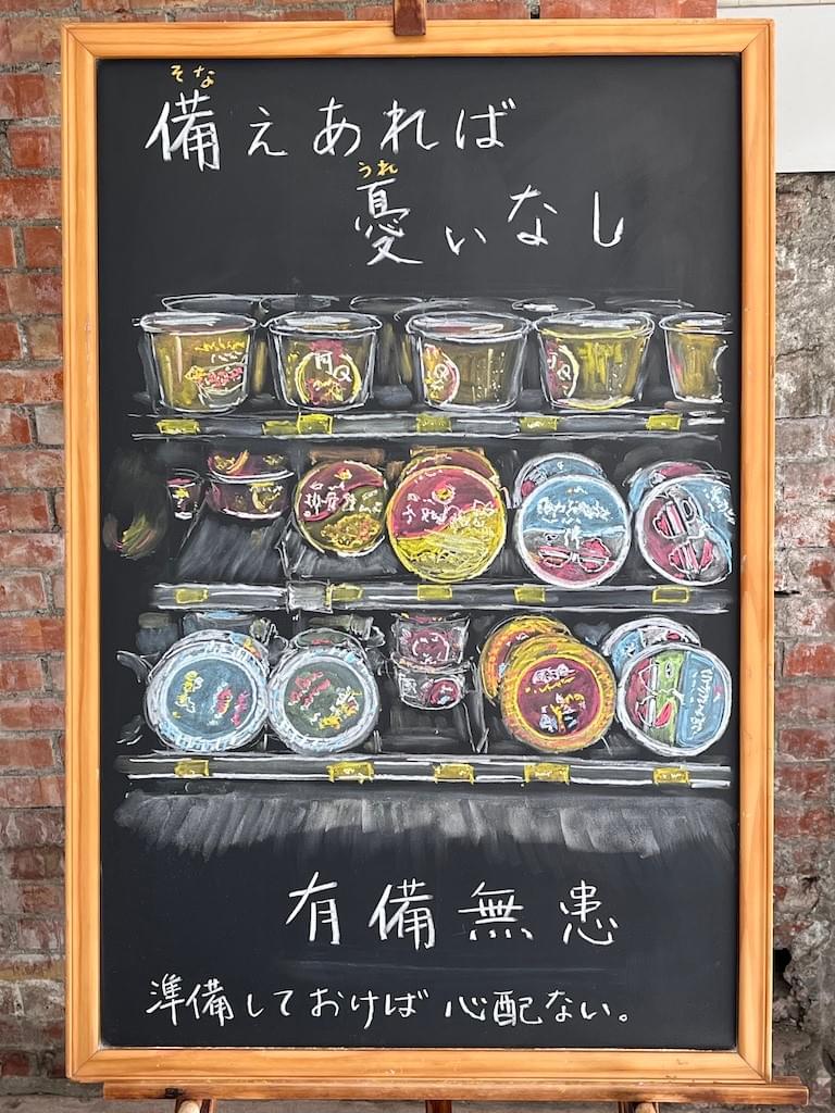小さい黒板にチョークで絵が描いてあります。日本語のことわざを紹介するための絵です。上部に横書きで「備えあれば憂いなし」とありまっす。中央に台湾のスーパーのカップ麺売り場の棚のイラストが描いて.あります。赤や黄色の華やかなパッケージのカップ麺が並んでいます。