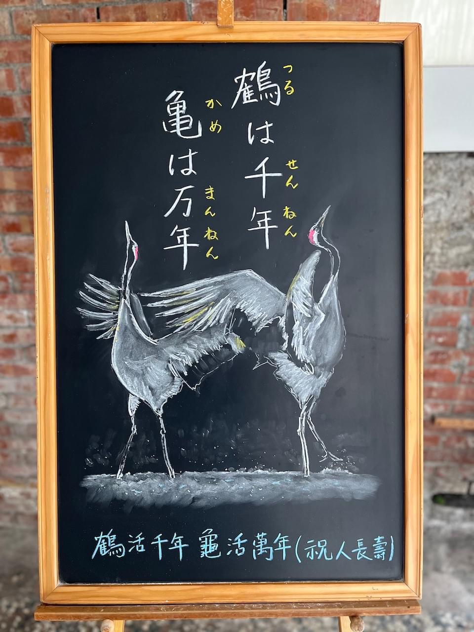 小さい黒板にチョークで絵が描いてあります。日本語のことわざ・慣用表現を紹介するための絵です。上部に縦書き二行で「鶴は千年亀は万年」とあり、下半分に二匹の鶴のイラストが描いてあります。雪の上で二羽の白い丹頂鶴が翼を広げ、首を伸ばし、求愛のダンスを踊っている様子です。