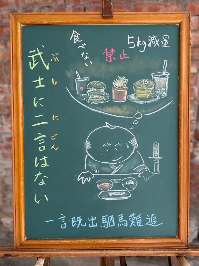 小さい黒板にチョークで絵が描いてあります。日本語のことわざを説明するための黒板です。 縦長の黒板で、左側に縦書き一行で「武士に二言はない」と書いてあります。 右側下に、武士と思われる男性のイラストが描いてあります。テーブルの上にはご飯、味噌汁と小さな焼き魚という質素な食事が置いてあります。男性は複雑な表情で料理を覗き込んでいます。男性の頭の中(吹き出しの中)には、高カロリーな食事のイラストがたくさん描いてあり、フライドポテト、ハンバーガー、パンケーキなどが並んでいます。その上に「禁止」「食べない」「５kg減量」などの文字も見えます。