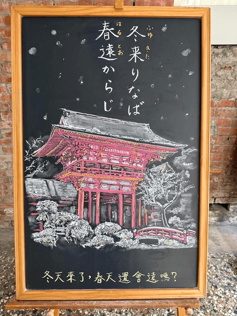 小さい黒板にチョークで絵が描いてあります。日本語のことわざを説明するための黒板です。 縦長の黒板で、上部に横書き二行で「冬来りなば／春遠からじ」と書いてあります。その下に赤い(朱色)柱の神社のイラストが描いてあります。雪が屋根に積もり、真っ白になっていて、付近の植物も雪をかぶって白く描いてあります。 ＊上賀茂神社の写真を見て描いたものです。