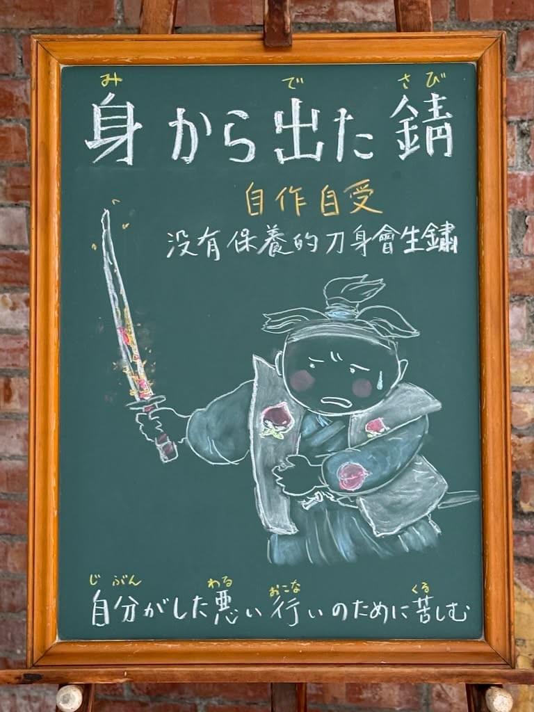 小さい黒板にチョークで絵が描いてあります。日本語のことわざを説明するための黒板です 縦長の黒板で、上部に横書き一行で「身から出た錆」と書いてあります。その下に、中文での説明「自作自受／沒有保養的刀身會生鏽」と書いてあります その下に桃太郎のような男の子のイラストが描いてあります。この男の子は、右手で刀を抜いたところですが、刀が錆びていて、悲しそうな顔をしています。