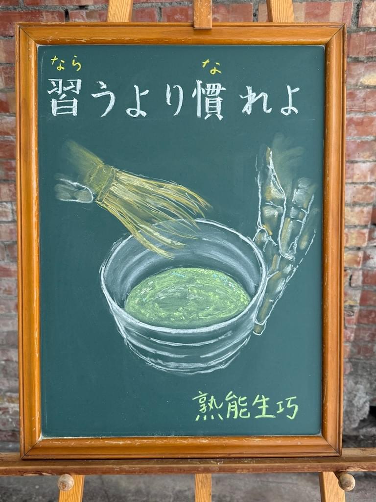 小さい黒板にチョークで絵が描いてあります。日本語のことわざを説明するための黒板です。 上部に横書き一行で「習うより慣れよ」と書いてあります。その下に、大きく茶道の茶碗のイラストが描いてあります。 白く丸い茶碗の中には点てられたばかりの抹茶が見え、茶碗の向かって右側に人の手が添えられていて、もう一方の手で茶筅が茶碗の中から引き上げられた場面です。