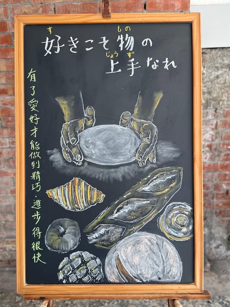 小さい黒板にチョークで絵が描いてあります。日本語のことわざを説明するための黒板です。 縦長の黒板で、横書き二行で「好きこそ物の／上手なれ」と書いてあります。 その下に、両手でパン生地をこねている様子のイラストが描いてあります。今は、丸まった白い生地を両手で包むようにしているところです。 その下に様々なパンのイラストが描いてあります。クロワッサン、バケット、ベーグル、メロンパンなどが見えます
