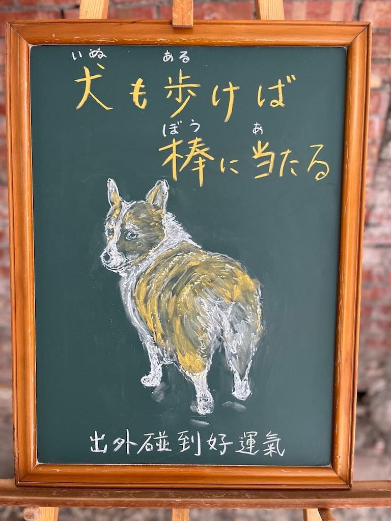 小さい黒板にチョークで絵が描いてあります。日本語のことわざを紹介するための絵です。上部に横書きで「犬も歩けば棒に当たる」とあります。中央にコーギー犬がこちらに後ろを見せた状態から、首を左へ回して振り向いているイラストが描いてあります。表情は「何か用？」とでも言いたげな興味なさそうな表情です。ちなみに、このコーギー犬の名前は「紅豆泥」です。
