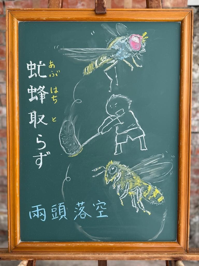 小さい黒板にチョークで絵が描いてあります。日本語のことわざを説明するための黒板です。 縦長の黒板で、左側に縦書きで「虻蜂取らず」と書いてあります。 右側上部に虻のイラスト、右側下部に蜂のイラストが大きく描いてあります。虻と蜂の間に、虫取り網を振り回す男の子のイラストが描いてあります。
