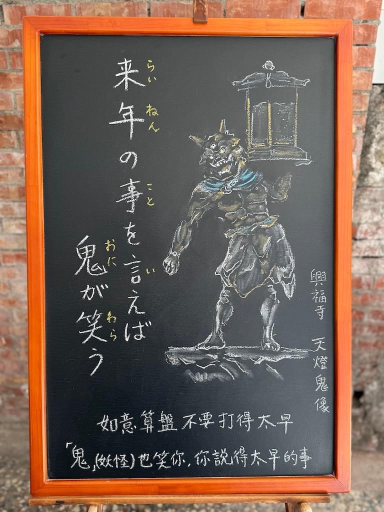 小さい黒板にチョークで絵が描いてあります。日本語のことわざ・慣用表現を紹介するための絵です。縦長の黒板で、左側に縦書き二行で「来年の事を言えば／鬼が笑う」と書いてあり、右側に興福寺にある彫刻、天燈鬼像のイラストが描いてあります。この像は、二本の角と3つの目を持つ鬼が、口を大きく開き、牙をむき出しにして、左肩に灯籠を載せて左手で支えている像です。下部に横書きで中国語の説明が書いてあります。如意算盤不要打得太早