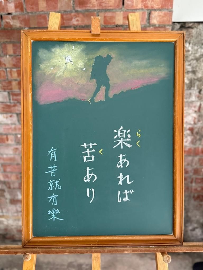 小さい黒板にチョークで絵が描いてあります。日本語のことわざ・慣用表現を紹介するための絵です。下半分に縦書き二行で「楽あれば／苦あり」と書いてあり、その左に一行で、この中国語「有苦就有樂」とあります。 上部には大きなリュックをかついだ登山者のイラストが描いてあります。左奥に黄色、白に輝く太陽があり、登山者の姿は影になっています。朝焼けで赤く染まった空を背景に黒い登山者の影が左から右に向けて坂道をのぼっている様子の絵です。
