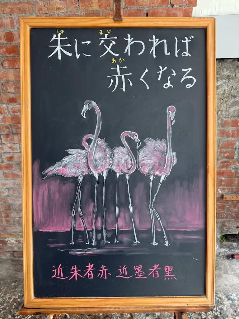 小さい黒板にチョークで絵が描いてあります。日本語のことわざを説明するための黒板です 縦長の黒板で、上部に横書き一行で「朱に交われば／赤くなる」と書いてあります。その下に、夕焼けの赤を背景に水辺に立っているフラミンゴ数羽のイラストが描いてあります。 下部にこのことわざの元になった中国語のことわざ「近朱者赤,近墨者黑」が書いてあります。