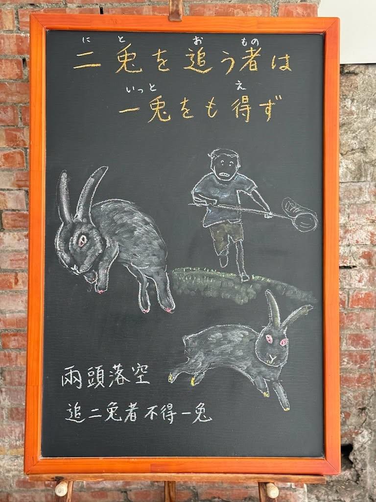 小さい黒板にチョークで絵が描いてあります。日本語のことわざを説明するための黒板です 縦長の黒板で、上部に横書き二行で「二兎を追う者は/一兎をも得ず」と書いてあり、その下に網を手にした男性が走って(手前に向かって)兎を追い掛けるイラストが描いてあります。その下に大きく（手前に向かって)逃げる二匹の白い兎のイラストが描いてあります