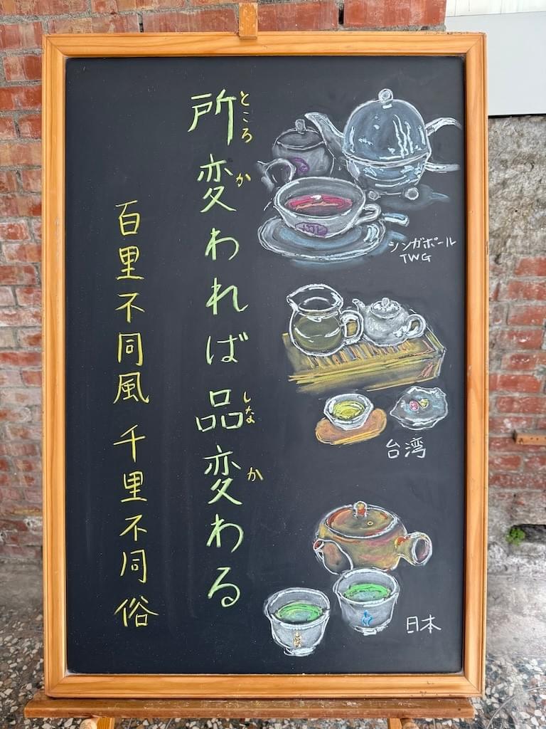 小さい黒板にチョークで絵が描いてあります。日本語のことわざ・慣用表現を紹介するための絵です。縦長の黒板で、左側に一行で「所変われば品変わる」と書いてあり、その左にこのことわざに相当する中国語の表現「百里不同風、千里不同俗」と書いてあります。 右側には、お茶の道具のイラストが三種類描いてあります。 上から紅茶のティーポットと、白いティーカップで、紅茶が注いであります。カップにはTWGという文字が見えます。シンガポールのTWGのカップです。 その下は、台湾のお茶セットにイラストです。木製の茶台の上にガラス製の茶海(ピッチャー)と白い陶器の小ぶりの茶壷(急須)のイラストが描いてあります。手前に茶杯があり、黄緑のお茶が注いであります。 一番下には日本茶の急須と湯呑2つのイラストが描いてあります。湯呑には緑茶が注いであります。