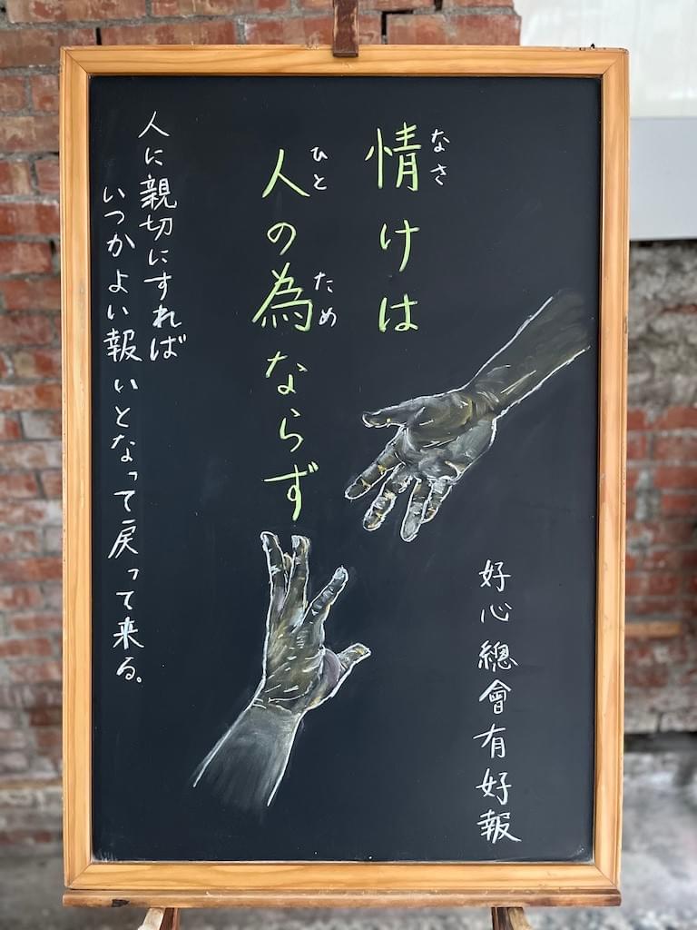 小さい黒板にチョークで絵が描いてあります。日本語のことわざ・慣用表現を紹介するための絵です。上部中央から縦書き２行で「情けは/人の為ならず」と書いてあります。人の手のイラストが描いてあります。下から助けを求めて伸ばした右手と、その手を掴もうと上から下に伸ばされた手。左脇に「人に親切にすれば/いつかよい報いとなって戻って来る」と書いてあります。