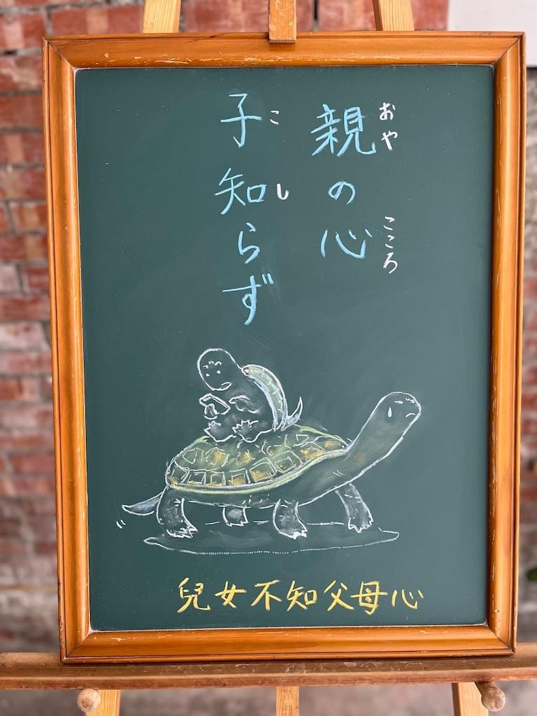 小さい黒板にチョークで絵が描いてあります。日本語のことわざ・慣用表現を紹介するための絵です。上部中央から縦書き２行で「親の心／子知らず」と書いてあります。その下に亀の親子のイラストが描いてあります。親亀の背中に乗った子亀はスマホに熱中している状態で、下の親亀はにがにがしい表情を見せています。