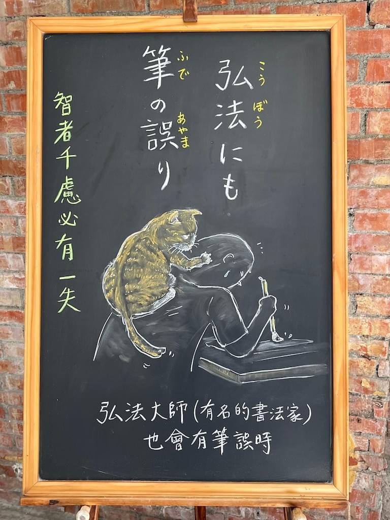 小さい黒板にチョークで絵が描いてあります。日本語のことわざ・慣用表現を紹介するための絵です。上部に縦書き二行で「弘法にも筆の誤り」と書いてあります。下部には、机に座った男性が筆を持って何か書こうとしているイラストが描いてあります。しかし、その男性の背中に、猫(茶トラ)が飛び乗って、男性の頭にちょっかいを出しており、字が書けない状況になっています。