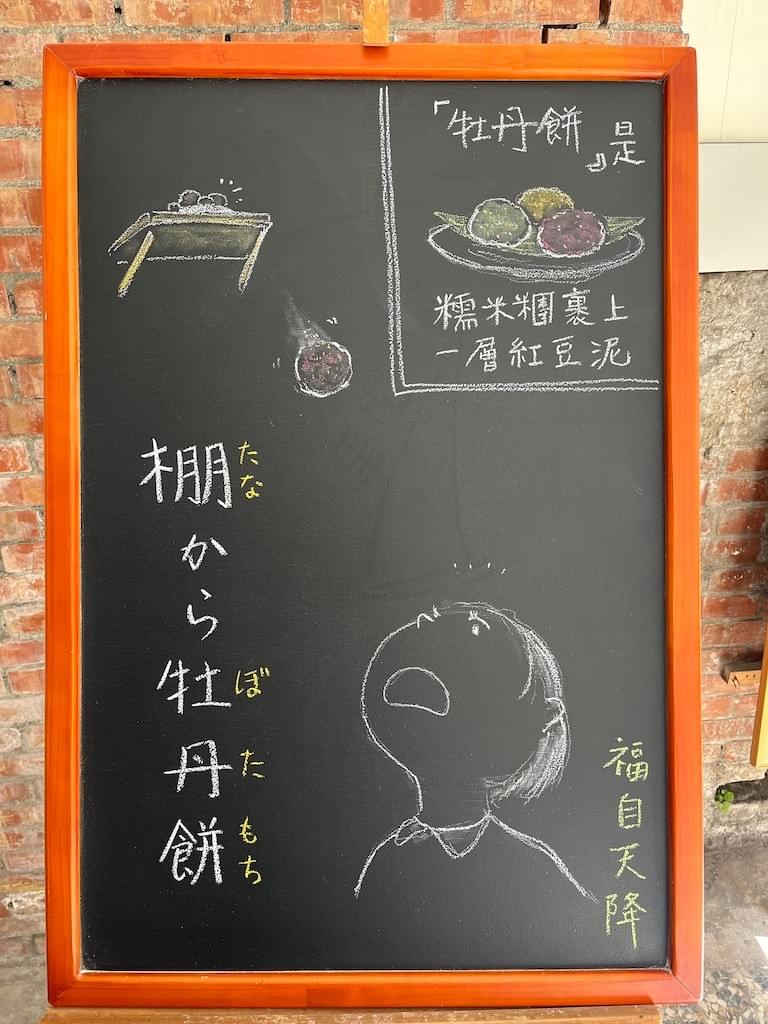 小さい黒板にチョークで絵が描いてあります。日本語のことわざ・慣用表現を紹介するための絵です。左下に縦書きで「棚から牡丹餅」と書いてあり、左上部の棚の上から丸い牡丹餅が一つ落ちてきています。右下に、その牡丹餅を驚いて見上げる人の顔のイラストが描いてあります。右上に皿の上に牡丹餅が3つのっているイラストと、中国語で、「牡丹餅」の説明があります。「牡丹餅」是糯米糰裹上ㄧ層紅豆泥」