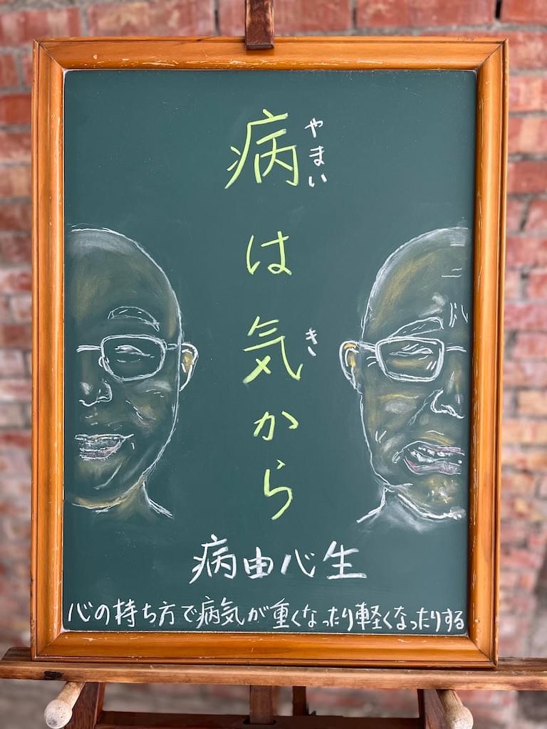 小さい黒板にチョークで絵が描いてあります。日本語のことわざ・慣用表現を紹介するための絵です。中央に縦書きで「病は気から」と書いてあり、その左右に人の顔のイラストが描いてあります。左側はメガネに、頭を剃った男性(犬山)が笑っている様子の顔半分、右は同じ男性が苦しげに顔を歪めている表情の絵が描いてあります