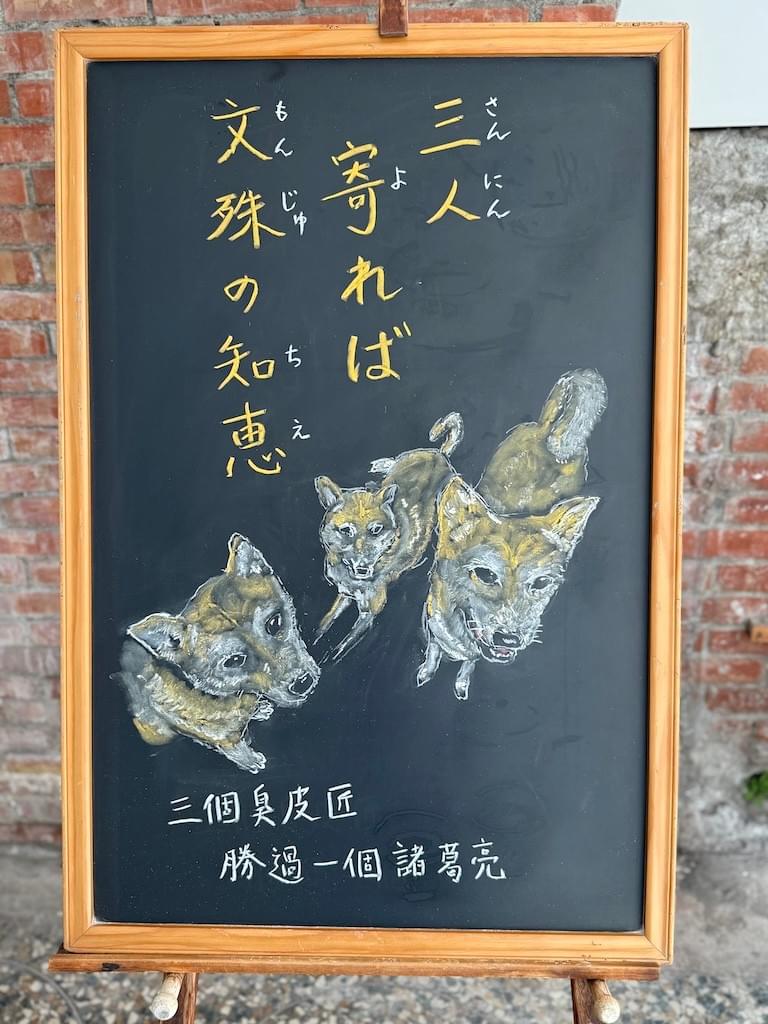 小さい黒板にチョークで絵が描いてあります。日本語のことわざ・慣用表現を紹介するための絵です。縦長の黒板の上部に縦書き三行で「三人／寄れば／文殊の知恵」と書いてあり、その下に三匹の柴犬のイラストが描いてあります。三匹を上から見下ろしたアングルで、三匹はそれぞれ上(こちら側)を見上げています。