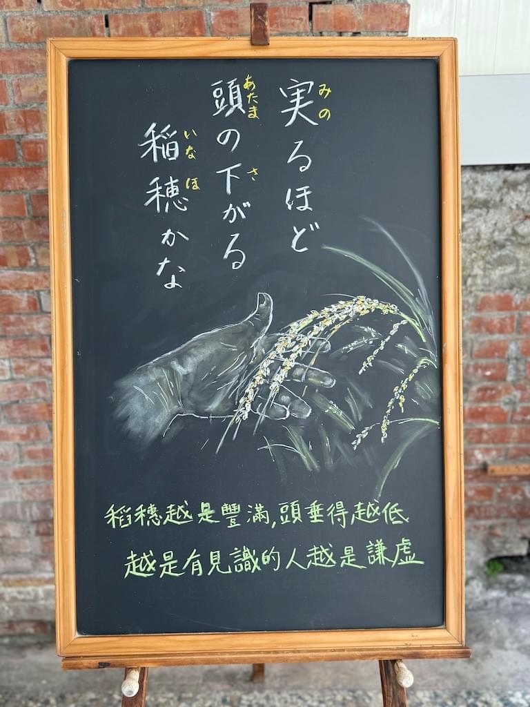 小さい黒板にチョークで絵が描いてあります。日本語のことわざ・慣用表現を紹介するための絵です。上部に三行縦書きで「実るほど/頭の下がる/稲穂かな」と書いてあります。その下に、左手で実った稲穂を触っている場面のイラストが描いてあります。