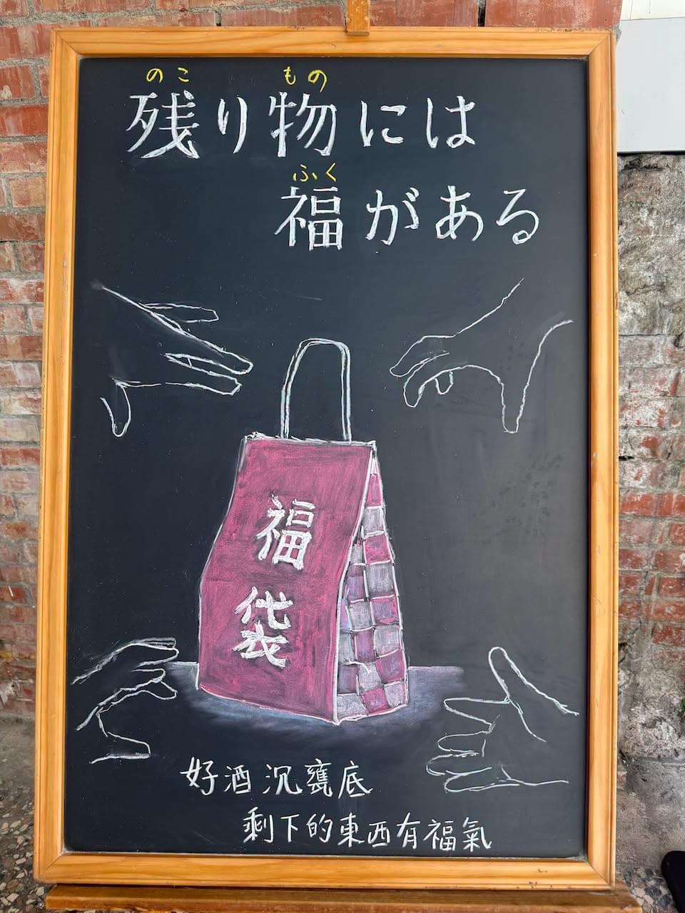 小さい黒板にチョークで絵が描いてあります。日本語のことわざを説明するための黒板です 縦長の黒板で、上部に横書き二行で「残り物には／福がある」と書いてあります。その下に赤い「福袋」のイラストが描いてあります。赤い紙製の手提げ袋に大きく白で「福袋」とあります。その袋を目指して、四方から人の手が伸びているイラストが描いてあります。