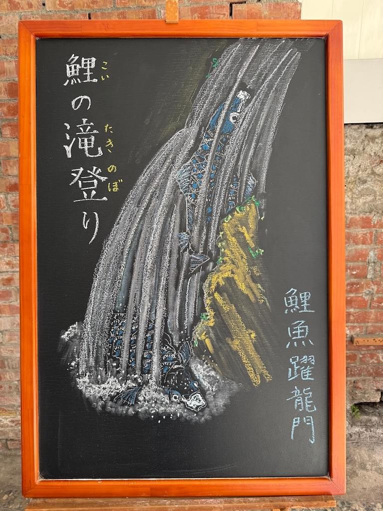 小さい黒板にチョークで絵が描いてあります。日本語のことわざを説明するための黒板です。 縦長の黒板で、中央にパイナップルを左手で持っているイラストが描いてあります。右がわに縦書き一行で「腹八分に医者いらず」左側に縦書き二行で「吃八分飽最合適／可以不用醫生」と書いてあります。下部に横書きで「満腹になるまで食べないで八分目で止めておけば病気をしない」と説明が書いてあります。