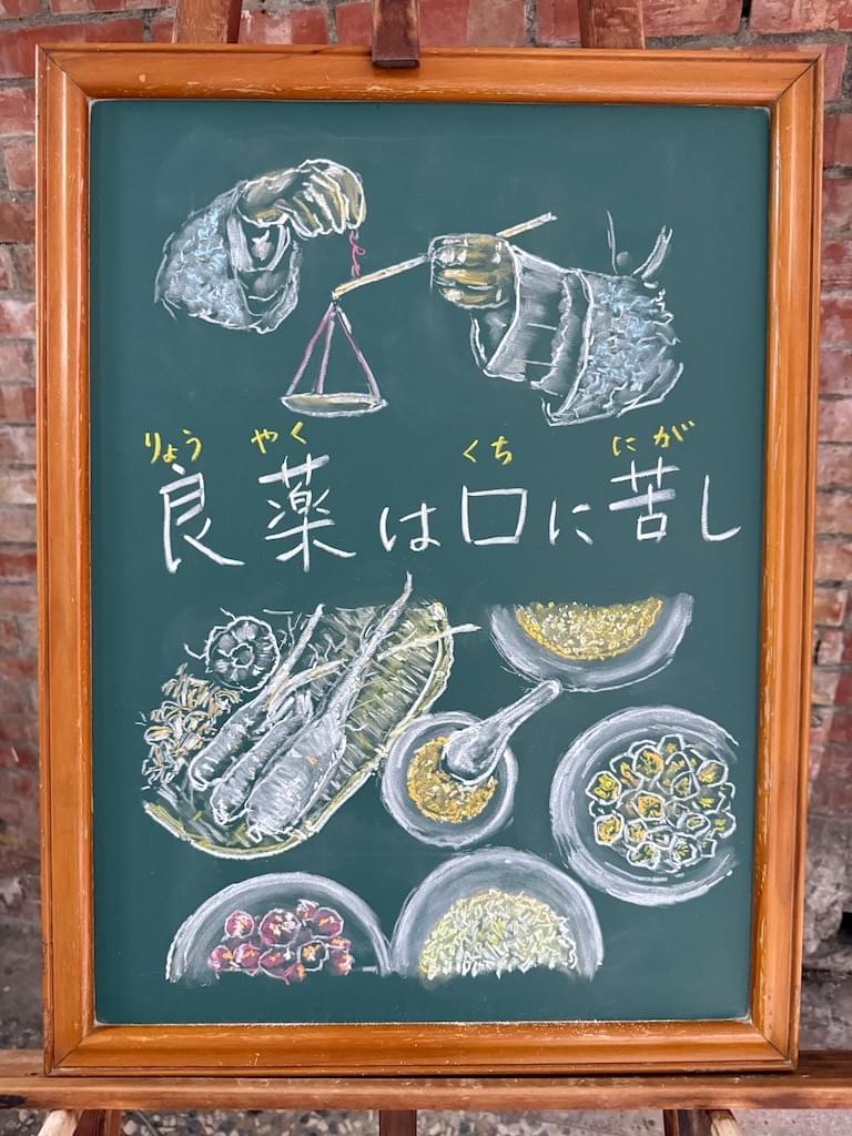 小さい黒板にチョークで絵が描いてあります。日本語のことわざ・慣用表現を紹介するための絵です。中央に横書きで「良薬は口に苦し」と書いてあります。その上部には、“唐装”中国式の長い袖のある服を着た人が両手で天秤を持ちながら薬の重さを量っている場面のイラストが描いてあります。腕の部分のみ。 下部には、テーブルの上に丸い小皿が並べられ、それぞれに様々な漢方薬の材料が入れてあるイラストが描いてあります。