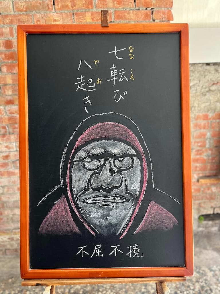 小さい黒板にチョークで絵が描いてあります。日本語のことわざ・慣用表現を紹介するための絵です。上部に縦書き２行で「七転び八起き」と書いてあります。その下に大きく「達磨」の顔のイラストが描いてあります。京都天龍寺にある「達磨図」を真似したものです。大きな目玉が斜め上をにらみ、口をぎゅっと結んでいます。