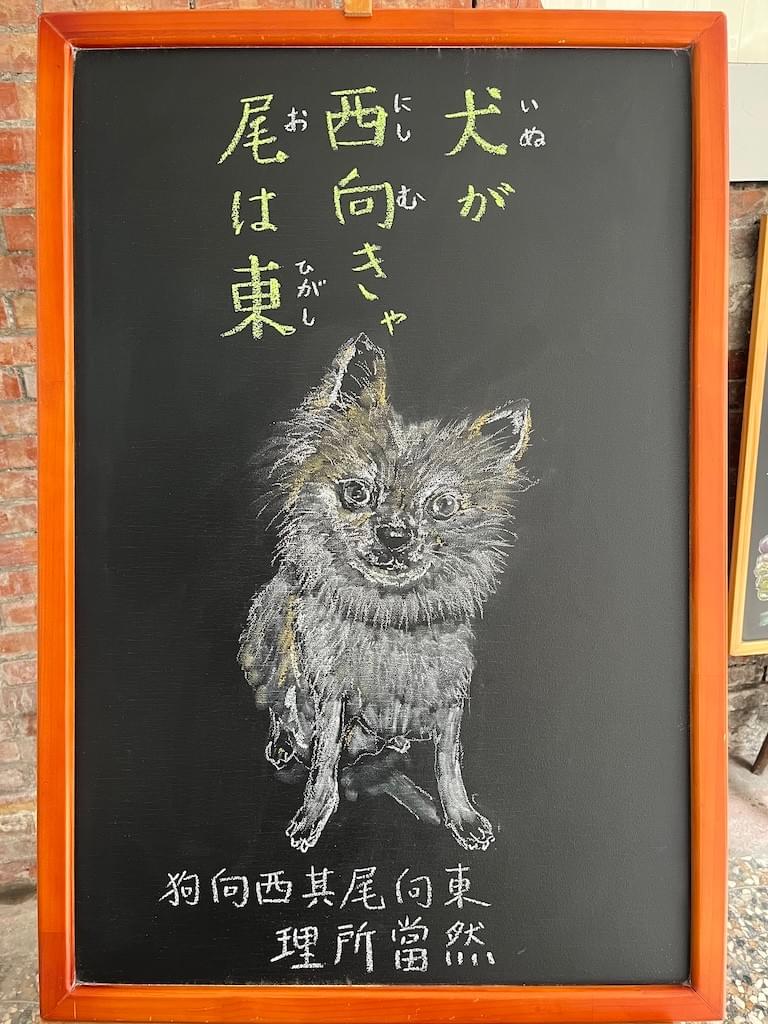 小さい黒板にチョークで絵が描いてあります。日本語のことわざ・慣用表現を紹介するための絵です。上部に横書き三行で「犬が西向きゃ尾は東」と書いてあります。その下に犬(ロングコートチワワ)が座っているイラストが描いてあります。白い毛がふさふさしていて、頭に少し茶色が入っています。