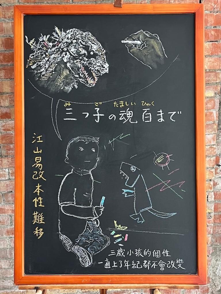 小さい黒板にチョークで絵が描いてあります。日本語のことわざを紹介するための絵です。中央やや上に横書きで「三つ子の魂百まで」と書いてあり、 下部には小さな男の子が壁にチョークで落書きをしているイラストが描いてあります。怪獣のようなものが口を開けている絵に見えます。 上部には、ゴジラのイラストと、チョークを持って絵を描いている右手のイラストが描いてあります。大人の手です。下の子どもが成長したあとの様子かもしれません。