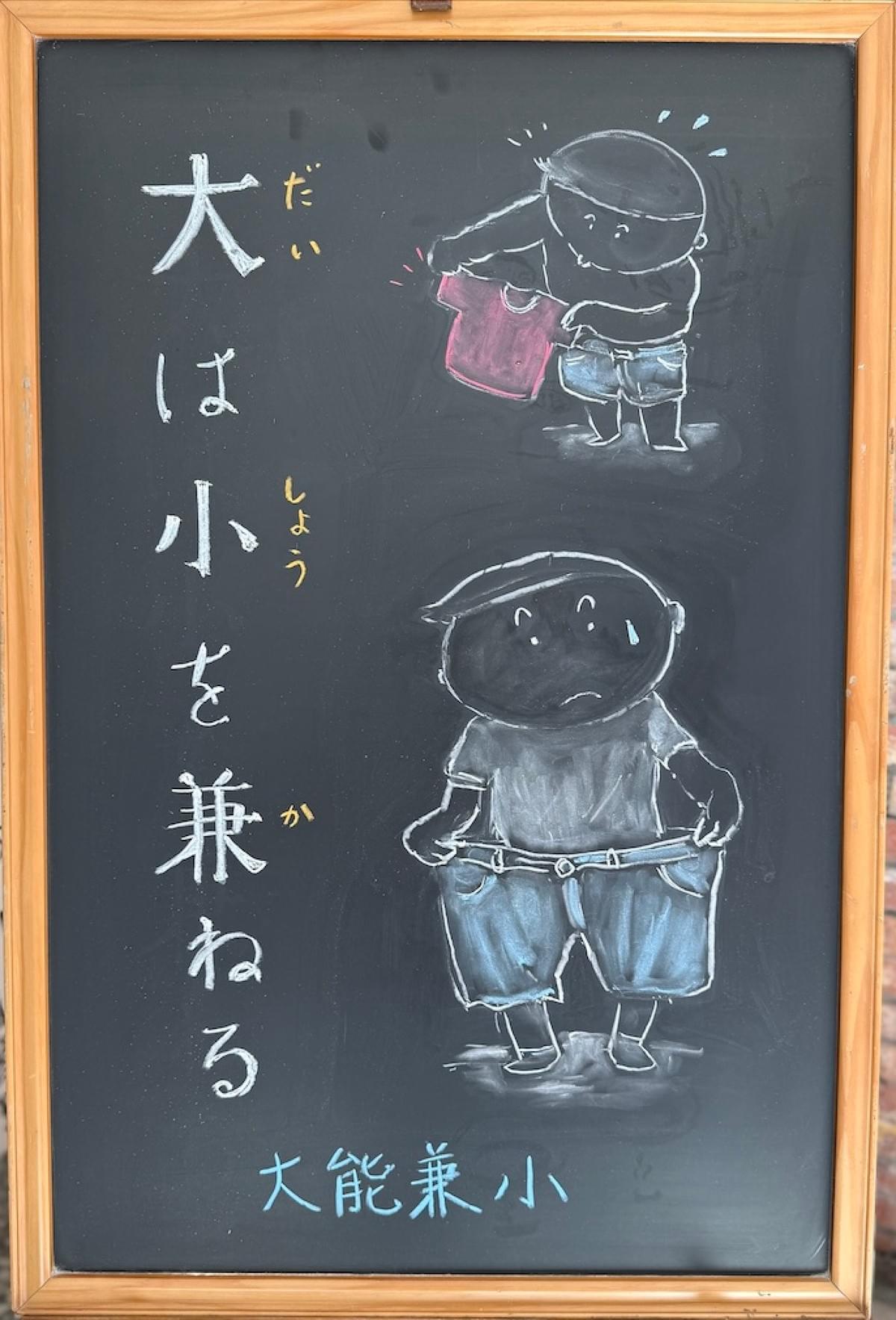 小さい黒板にチョークで絵が描いてあります。日本語のことわざを説明するための黒板です。 縦長の黒板で、左側に縦書き一行で「大は小を兼ねる」と書いてあります。 その左側に、男の子のイラストが描いてあります。 下部の男の子は、かなり大きな半ズボンをはこうとして、もてあましている様子。困惑している表情です。 上部の男の子は、かなり小さなTシャツを両手で持って、どうしたものかと考えている様子。