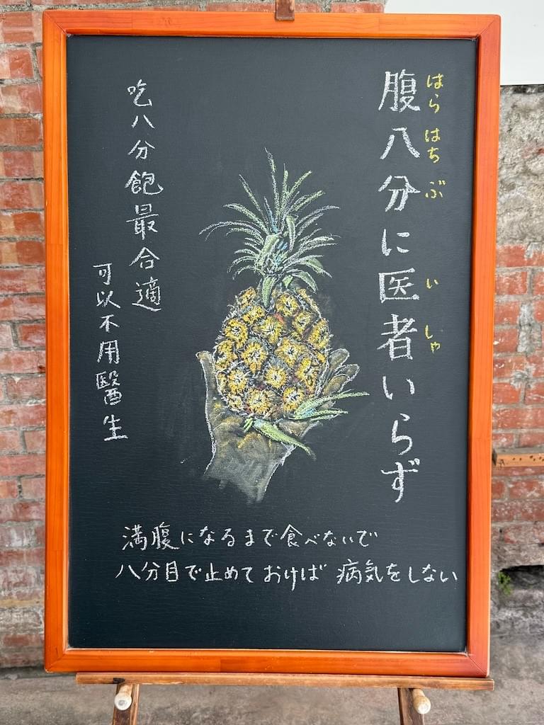 小さい黒板にチョークで絵が描いてあります。日本語のことわざを説明するための黒板です。 縦長の黒板で、中央にパイナップルを左手で持っているイラストが描いてあります。右がわに縦書き一行で「腹八分に医者いらず」左側に縦書き二行で「吃八分飽最合適／可以不用醫生」と書いてあります。下部に横書きで「満腹になるまで食べないで八分目で止めておけば病気をしない」と説明が書いてあります。
