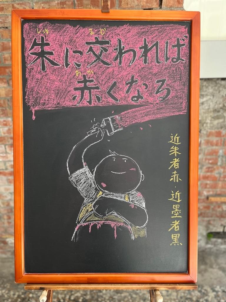 小さい黒板にチョークで絵が描いてあります。日本語のことわざ・慣用表現を紹介するための絵です。上部に横書き２行で「朱に交われば／赤くなる」と書いてあります。背景は赤で文字の部分に色はなく黒板の地の色が見えています。 下部に赤いペンキを塗っている男の子のイラストが描いてあります。右手に刷毛を持って上部を勢いよく塗っていて、もう一方の手でペンキの缶を前に持っています。