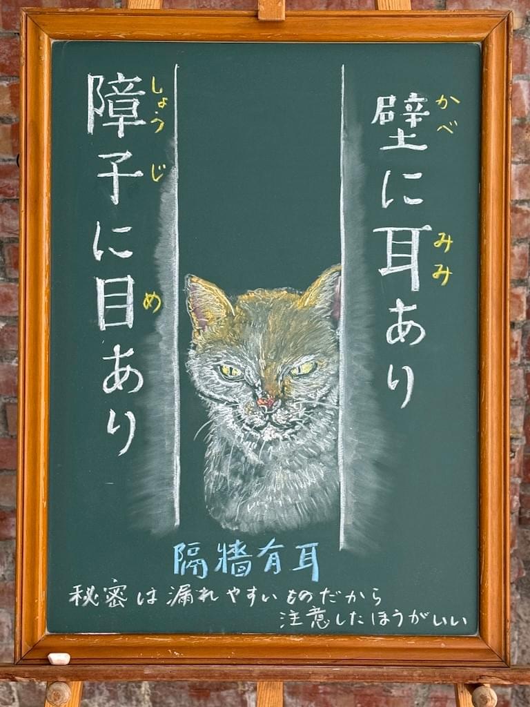 小さい黒板にチョークで絵が描いてあります。日本語のことわざを説明するための黒板です。 縦長の黒板で、中央に壁と壁の間から、こちらを覗いている猫のイラストが描いてあります。口を結んでこちらをじっと睨んでいるような、ちょっと怖い感じの顔です。その左右両側に分かれて、ことわざが縦書きで書いてあります。右に「壁に耳あり」左に「障子に目あり」