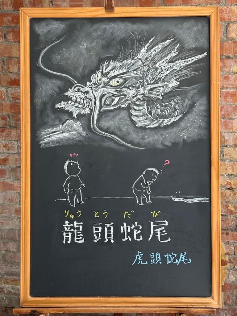 小さい黒板にチョークで絵が描いてあります。日本語のことわざを説明するための黒板です。 縦長の黒板で、上半分に大きな龍の顔のイラストが描いてあります。顔は横向きで長いヒゲがあり、剥き出しの歯を見せ、目玉はこちらをぎょろりと睨んでいる様子です（天龍寺の雲龍図の写真を見て描きました) その下に、人のイラストが描かれていて、一人は上の龍を見上げて、びっくりしている様子、もう人は足元に見える、細い蛇の尻尾を見て、「ん？」と考えている様子です。 その下に横書き一行で「龍頭蛇尾」と書いてあります。その下に、このことわざに相当する中国語の「虎頭蛇尾」が書いてあります。