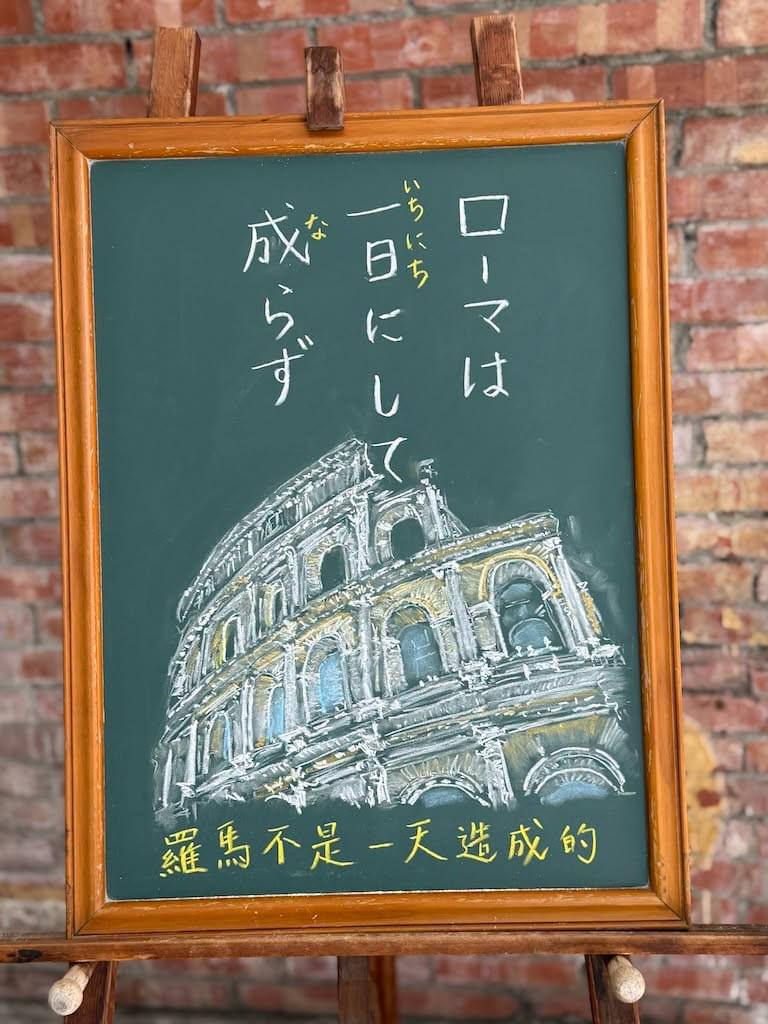 小さい黒板にチョークで絵が描いてあります。日本語のことわざを説明するための黒板です 縦長の黒板で、上部に縦書き三行で「ローマは/一日にして/成らず」と書いてあります。その下に、現代ローマに残るコロッセウムの壁のイラストが描いてあります。