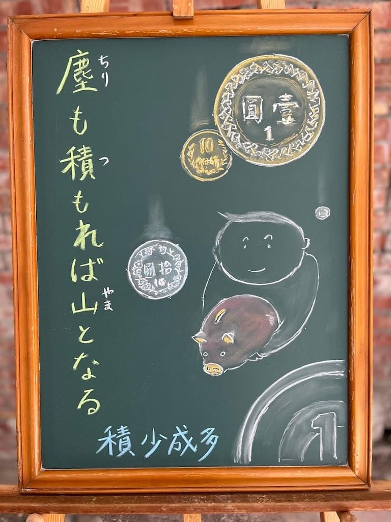 小さい黒板にチョークで絵が描いてあります。日本語のことわざ・慣用表現を紹介するための絵です。左に縦書き一行で「塵も積もれば山となる」と書いてあります。右側に男の子がピンクの豚の貯金箱を重そうに抱えているイラストが描いてあります。また、背景や手前の部分にたくさんのコインのイラストが描いてあります。台湾の1元や、10元、日本の1円や１０円玉の絵です。