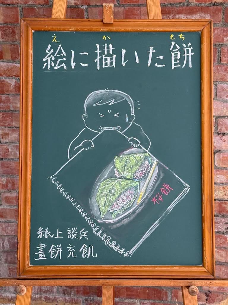 小さい黒板にチョークで絵が描いてあります。日本語のことわざを説明するための黒板です 縦長の黒板で、上部に横書き一行で「二絵に描いた餅」と書いてあり、その下にスケッチブックの絵をかんでいる男の子のイラストが描いてあります。スケッチブックには、お皿に載った桜餅２個のイラストが描いてあります。