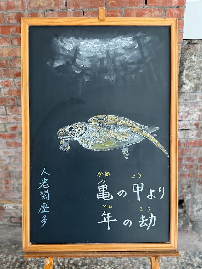 小さい黒板にチョークで絵が描いてあります。日本語のことわざ・慣用表現を紹介するための絵です。縦長の黒板で中央に海の中を泳ぐウミガメのイラストが描いてあります。その上に水面らしき部分が光を浴びて白くなっています。ウミガメの下に横書き二行で「亀の甲より/年の劫」と書いてあります。左下に、このことわざの説明にあたる中国語が縦書きで小さめに書いてあります「人老閱歷多」