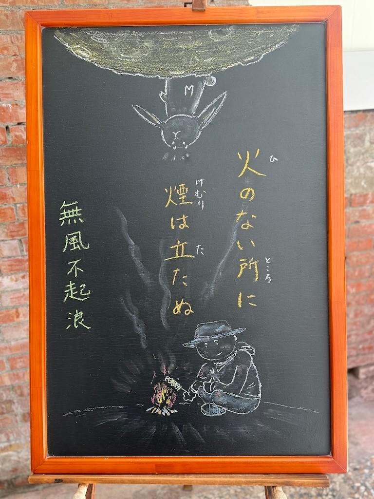 小さい黒板にチョークで絵が描いてあります。日本語のことわざ・慣用表現を紹介するための絵です。縦長の黒板で中央に二行で「火のない所に／煙は立たぬ」と書いてあり、下部にキャンプで焚き火をしている人のイラストが描いてあります。帽子を被り、長袖のシャツで地面に直接座り込んでいます。手に串に刺した肉(のような物)を持っていて焚き火の上にかざしています。そこから、白い煙が上部に上がっています。上部には月とおぼしき球体の一部があり、そこに立ったウサギが下から上がってきた煙に気づいた様子のイラストが描いてあります。