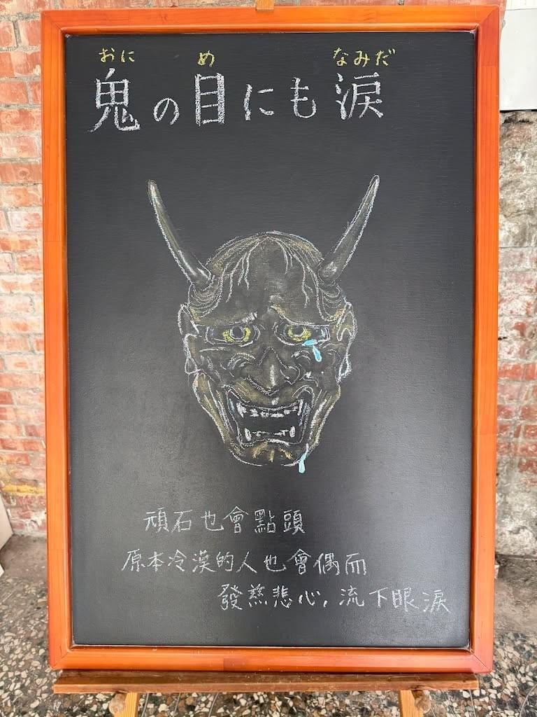 小さい黒板にチョークで絵が描いてあります。日本語のことわざを説明するための黒板です 縦長の黒板で、上部に横書き一行で「鬼の目にも涙」と書いてあります。 その下に能面、般若の面のイラストが描いてあります。面の左目から涙がこぼれ、あごの辺りまで落ちています。 その下に、この日本語のことわざの説明が中文で書いてあります 頑石也會點頭、原本冷漠的人也會偶而發慈悲心、流下眼淚
