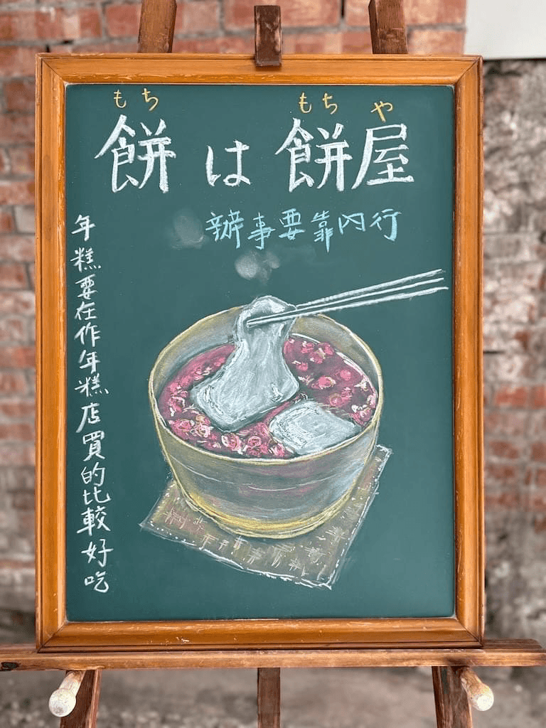 小さい黒板にチョークで絵が描いてあります。日本語のことわざを説明するための黒板です。 縦長の黒板で、上部に横書き一行で「餅は餅屋」と書いてあります。下部に御椀に入ったおしるこのイラストが描いてあります。 白い餅が二切れ浮かんでいて、箸でそのひとつを持ち上げている様子です。