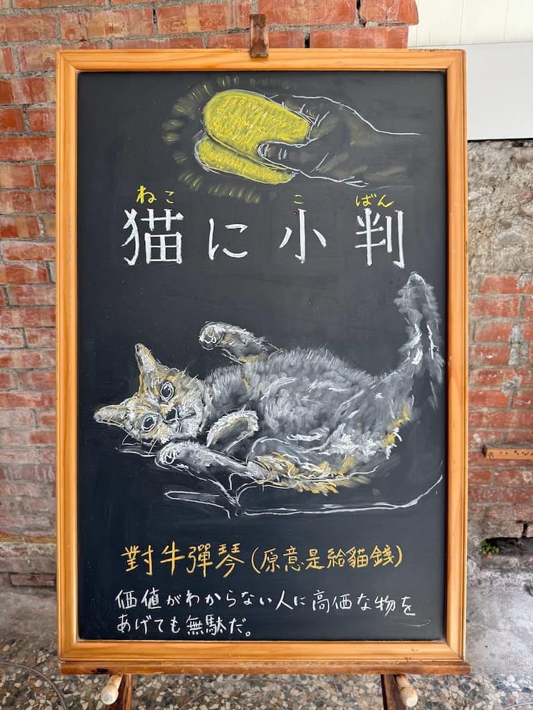 小さい黒板にチョークで絵が描いてあります。日本語のことわざを説明するための黒板です 縦長の黒板で、上部に小判を二枚もっている人の右手のイラスト、 その下に横書き一行で「猫に小判」とあり、 その下に、寝っ転がって、おなかを見せた状態で、顔はこちらがわを見ている猫にイラストが描いてあります。